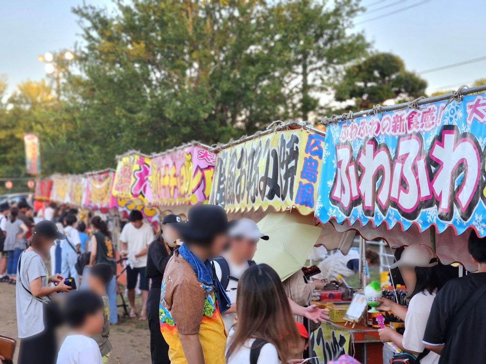 夕暮れの公園に並ぶ屋台に人が集まり、たこ焼きやお好み焼き、ふわふわ系スイーツなどの看板が並ぶ様子