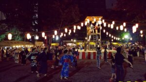 夜、木製の二層櫓を囲んで放射状に提灯が吊られ、その下で浴衣姿の人々が輪になって踊っている