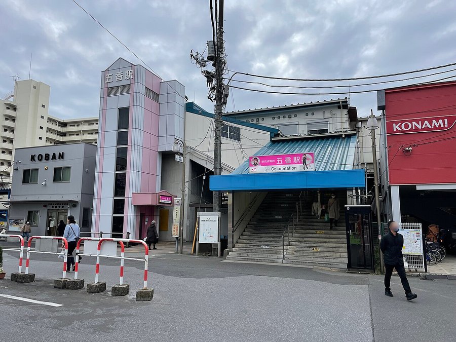 曇天の駅前。ピンクと白のエレベーター塔屋に「五香駅」の縦書き、青い庇の付いた階段入口、左手前に交番が見える