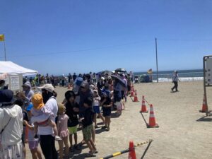 快晴の砂浜で、カラーコーンとバーで仕切られた列に親子連れが並んでいる。奥の波打ち際には赤と黄のフラッグが立つ