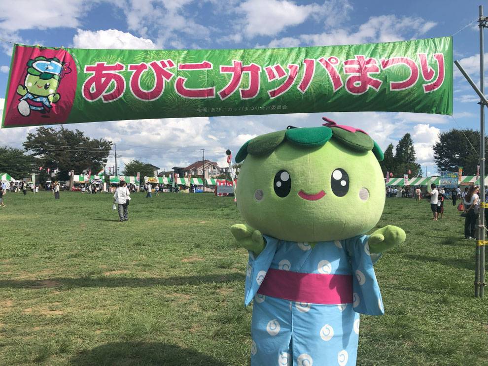 晴れた日中の芝生広場に「あびこカッパまつり」と書かれた緑と赤の大きな横断幕が張られ、その下で水色の浴衣を着たカッパの着ぐるみが両手を広げて立っている。背後には白と緑のストライプのテント屋台が並び、来場者が歩いている