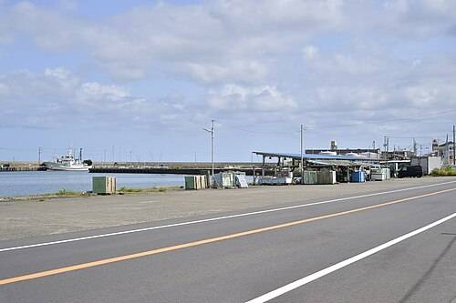 昼間の漁港。手前にオレンジのセンターラインが引かれた舗装道路が横切り、その向こうに緑色のコンテナや小屋が並び、左奥の岸壁に白い漁船が1隻停まっている