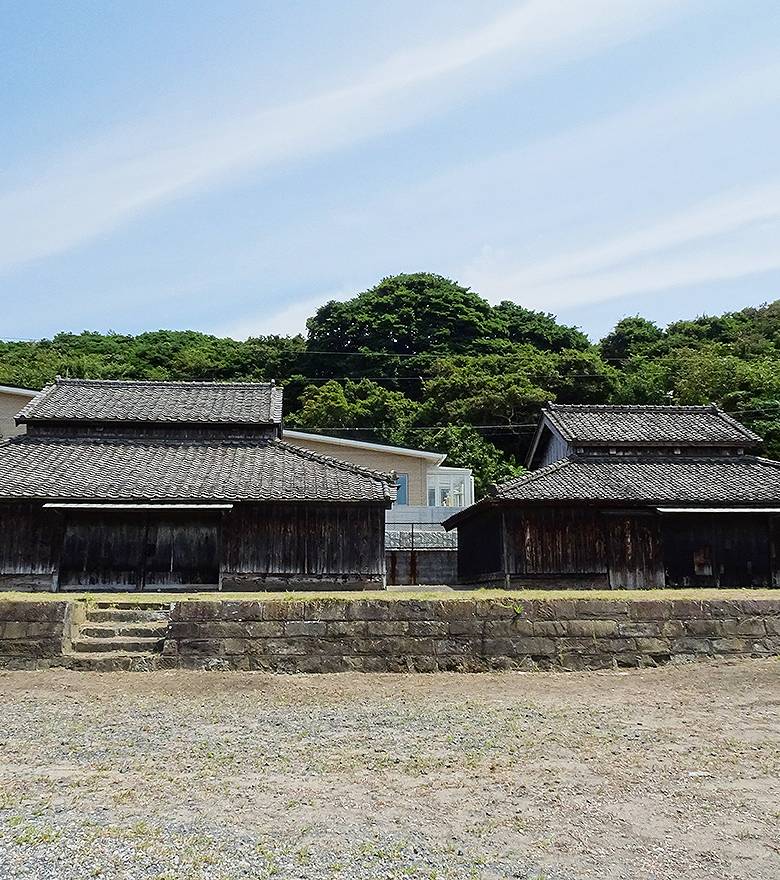 黒く焼けた縦板張りの平屋の建物が2棟並んでいる。瓦屋根の庇が深く張り出し、窓はほとんど見えない。手前に石積みの擁壁と石段、その前に砂利の空き地。背景は緑の丘と夏空