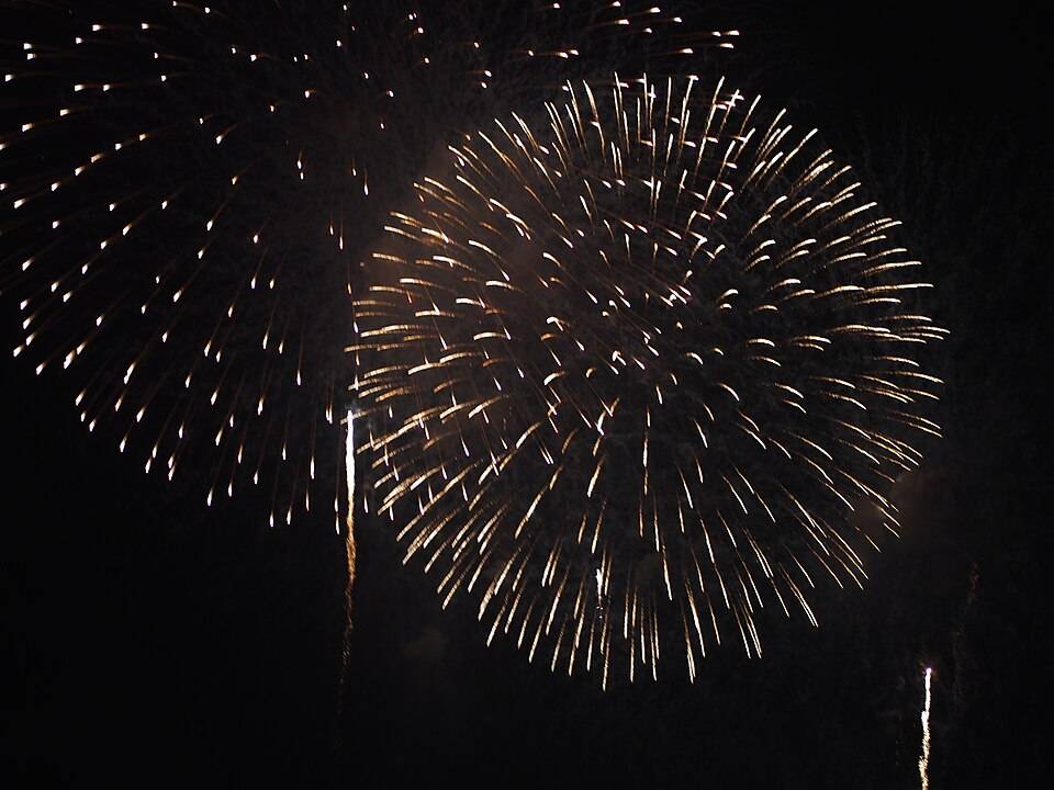 真っ暗な夜空に、金色の火の粉が大きく円形に広がる打ち上げ花火が2発ひらいている。下から尾を引いて上がる光の筋も見える