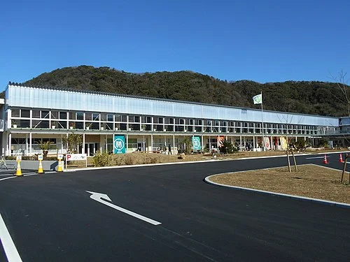 小学校の校舎をそのまま使った横長2階建ての白い建物。1階には店舗の幟が並び、手前は舗装された駐車場と車道、背後に低い山が見える