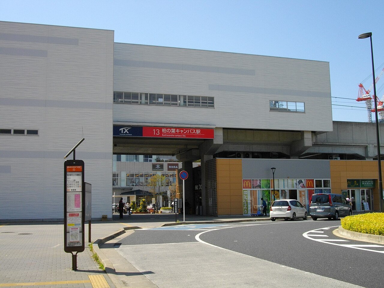 つくばエクスプレス柏の葉キャンパス駅の西口。高架下の壁に赤い駅名看板「13 柏の葉キャンパス駅 Kashiwanoha-campus Sta.」があり、手前のロータリーにバス停の時刻表ポール、右手にマクドナルドなどの店舗、奥に建設中のクレーンと青空