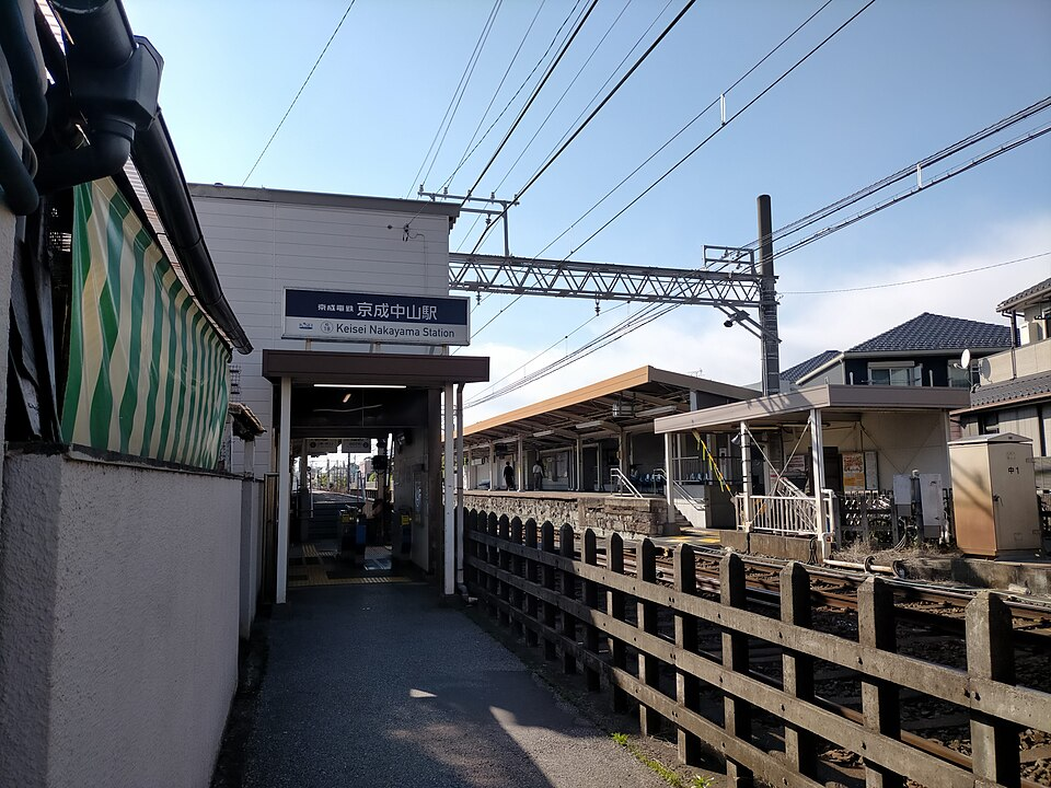 京成中山駅の小さな駅舎と、線路沿いに続く細い通路。ホームの屋根と架線柱が見える