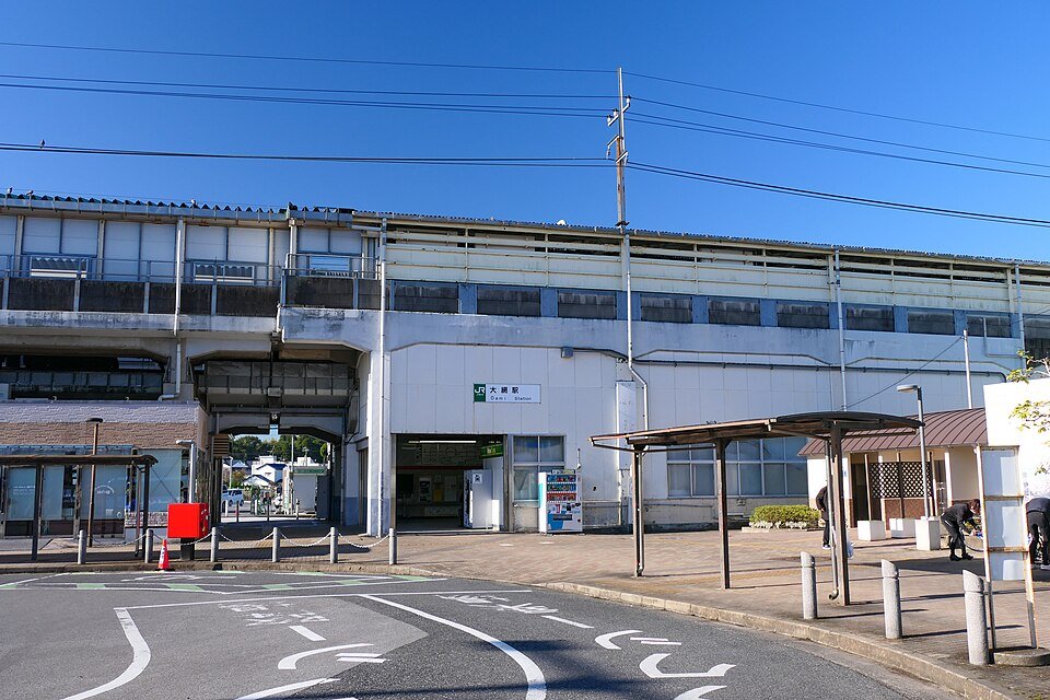 JR大網駅の駅舎外観。高架のホーム下に「大網駅」の駅名看板と改札口があり、手前はロータリーの車道。青空が広がっている