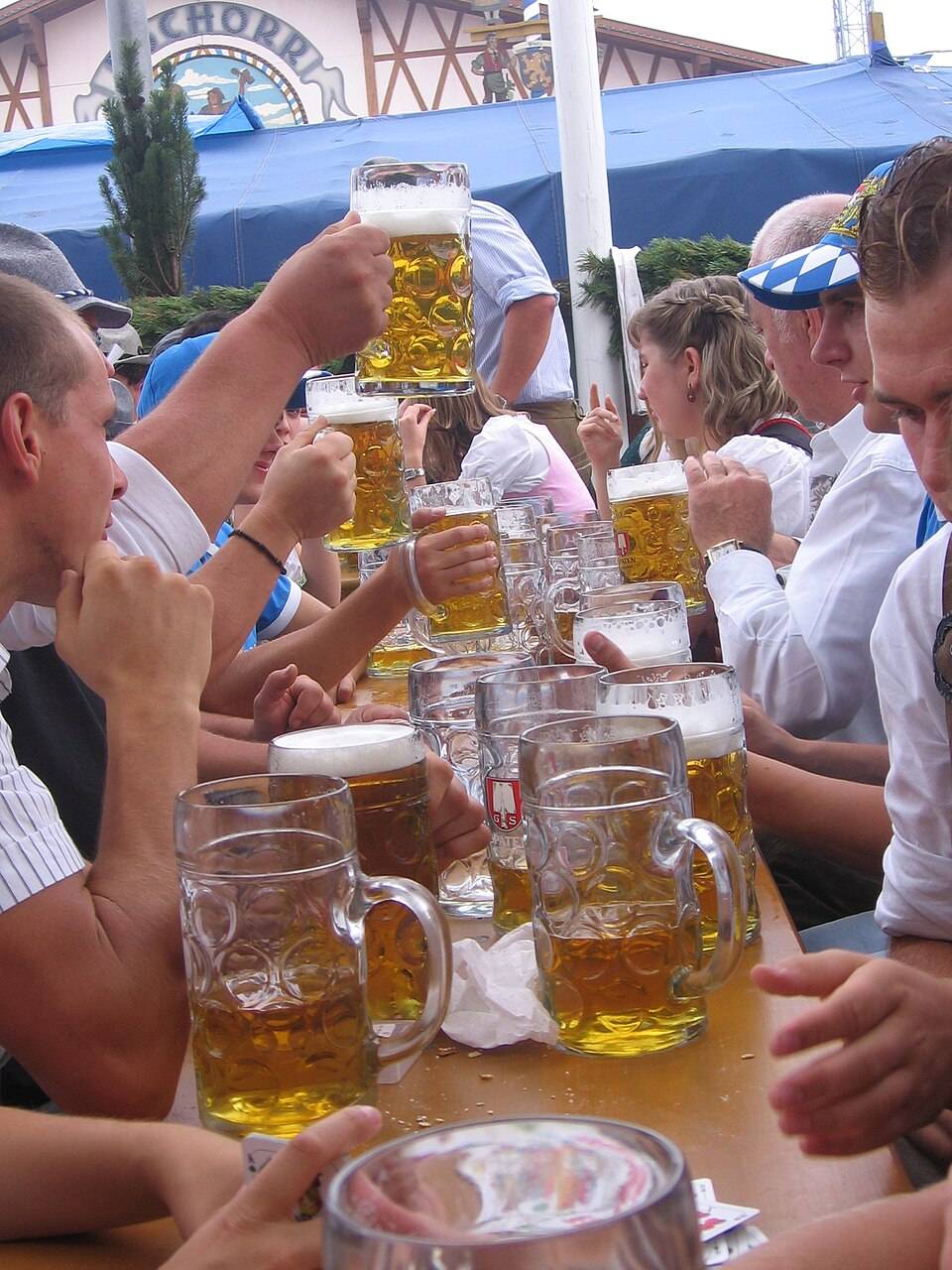 ビアテントの前で、大きなガラスの1リットルジョッキになみなみと注がれた金色のビールを、大勢の人が手に持って掲げている。テーブルの上にも同じ大ジョッキがいくつも並ぶ