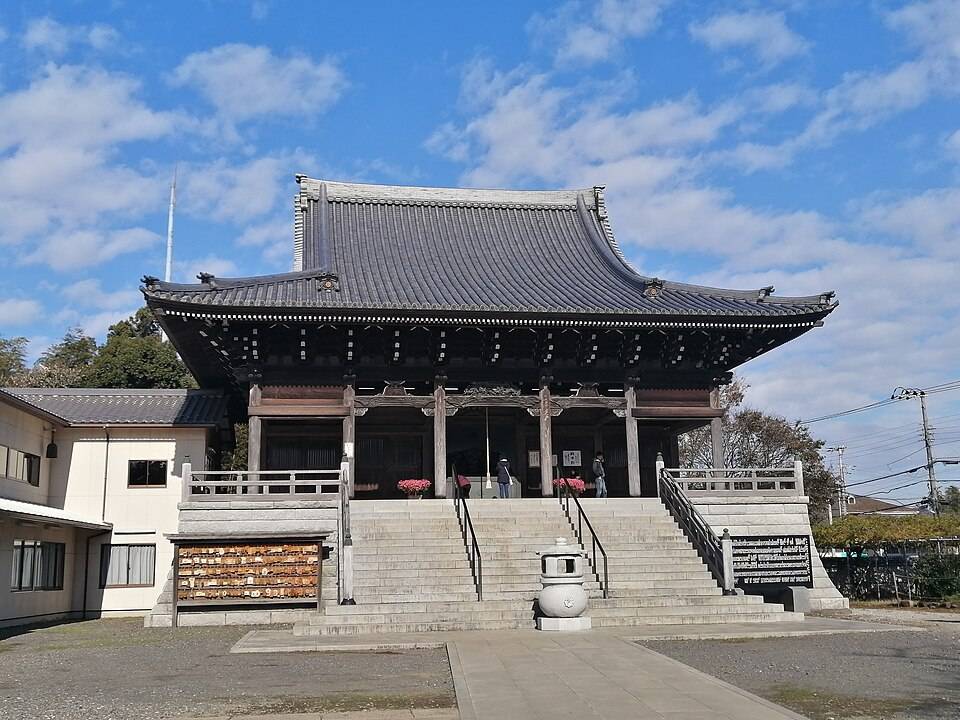 青空の下、正面から見た金蔵寺の本堂。反り屋根の瓦葺きで、正面に長い石段が伸び、左手に絵馬掛けと石灯籠がある