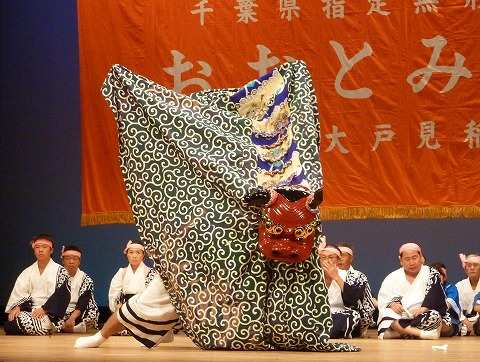 唐草模様の胴幕に二人が入った獅子が赤い獅子頭を高く掲げて舞い、白装束に鉢巻き姿の囃子方が両脇に座っている