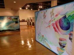 屋内の展示室。木目の床に照明が反射し、左奥の壁に風景画、右手前に淡い色づかいの大きな抽象画のパネルが立てられている