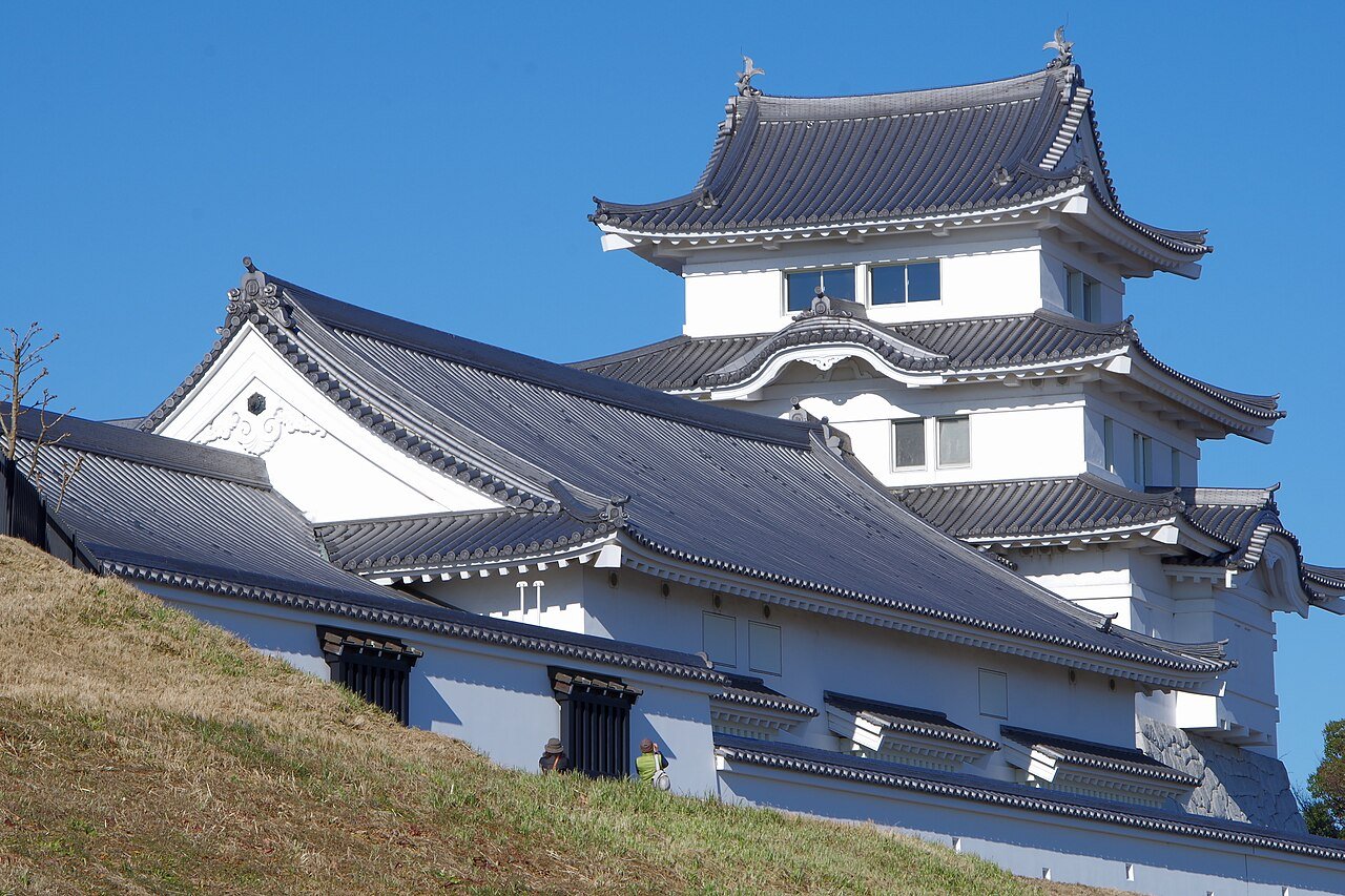 青空を背に建つ白い天守。関宿城博物館の建物で、黒瓦の入母屋屋根を重ね、最上階の白壁に窓が並ぶ。手前は枯れ草の土手で、階段の下に見学者の姿がある