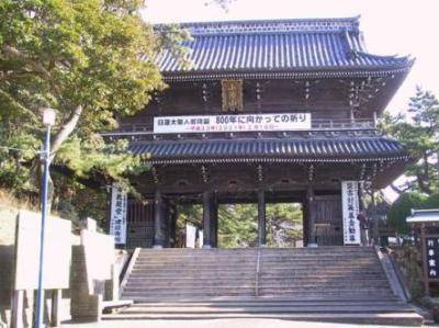 昼間の誕生寺の仁王門。石段の上に建つ二層の木造門で、正面に横断幕が張られている