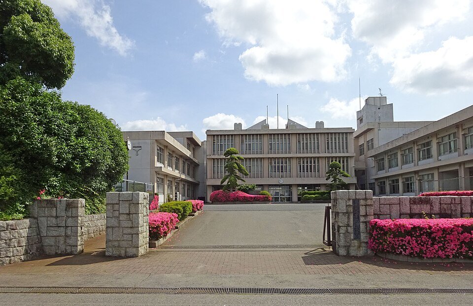 千葉県立富里高等学校の正門。石積みの門柱の奥に3階建ての校舎が建ち、手前の花壇に濃いピンクのツツジが咲いている。空は青く雲が浮かぶ