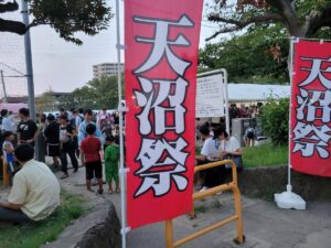 夕方の公園に立つ、赤地に白抜きで「天沼祭」と大書されたのぼり旗2本。奥に浴衣姿の子どもや家族連れ、テント、飲食する来場者が見える