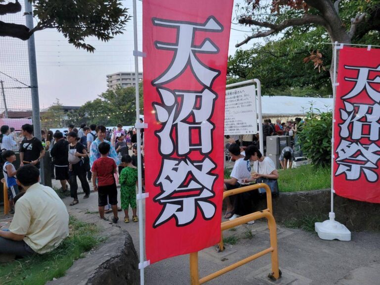夕方の公園に立つ、赤地に白抜きで「天沼祭」と大書されたのぼり旗2本。奥に浴衣姿の子どもや家族連れ、テント、飲食する来場者が見える