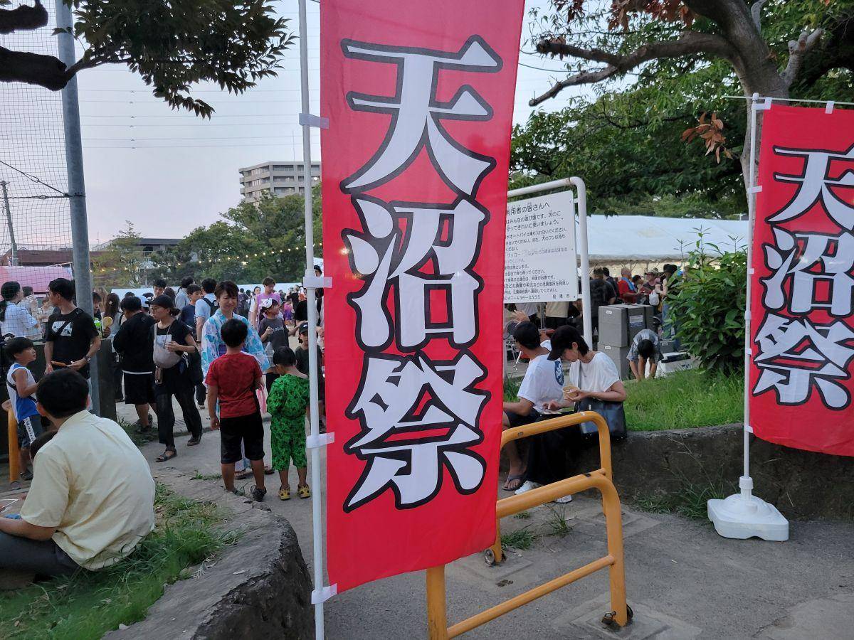 夕方の公園に立つ、赤地に白抜きで「天沼祭」と大書されたのぼり旗2本。奥に浴衣姿の子どもや家族連れ、テント、飲食する来場者が見える