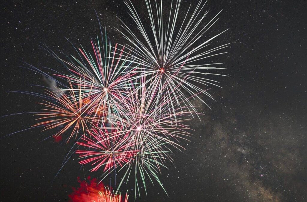 星が散る夜空に、白・赤・緑の大きな花火が3発ほど同時に開いている。右奥にうっすらと天の川がかかり、画面下からは赤い光が立ちのぼっている