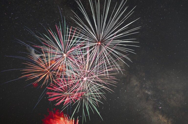 星が散る夜空に、白・赤・緑の大きな花火が3発ほど同時に開いている。右奥にうっすらと天の川がかかり、画面下からは赤い光が立ちのぼっている