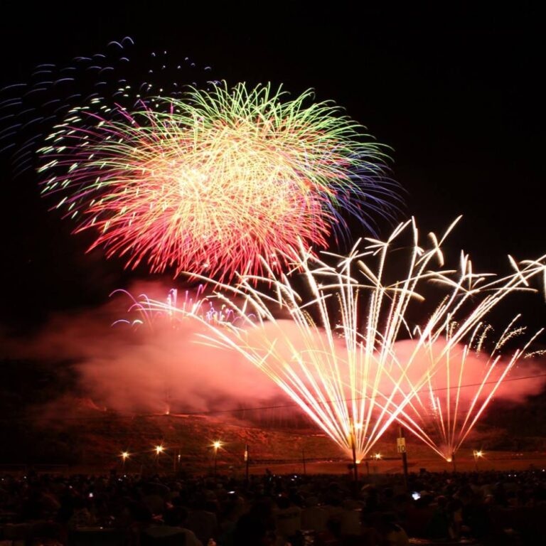 夜空に大玉の花火と扇状に広がるスターマインが同時に開き、手前に観客席のシルエットと会場の照明が並んでいる