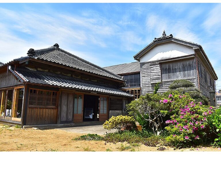 旧西廣家住宅の主屋。左手に瓦屋根の平屋が建ち、格子戸と障子が並んで玄関の引き戸が開いている。右奥には白漆喰の妻壁を持つ板張りの2階建て。手前の庭にピンクのツツジが咲き、青空が広がっている