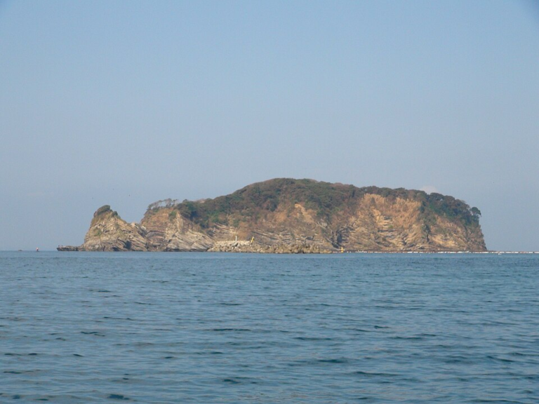 穏やかな海に浮かぶ無人島。岩肌がむき出しの崖の上に低い木々が茂り、左端に小さくとがった岩が立っている