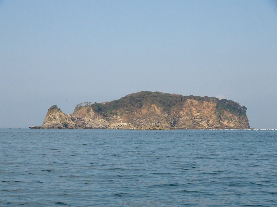 穏やかな海に浮かぶ無人島。岩肌がむき出しの崖の上に低い木々が茂り、左端に小さくとがった岩が立っている