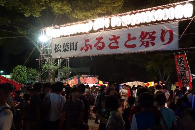 夜の松葉町ふるさと祭りの入口。ゲートに「松葉町ふるさと祭り」と大きく書かれた白い横断幕が張られ、上には紅白の飾りと提灯がずらりと並ぶ。奥に大判焼き・焼きそば・たこ焼きなどの露店と「焼きそば」ののぼり、手前には浴衣や私服の大勢の来場者が行き交う
