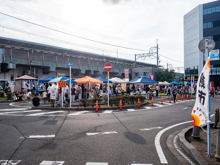 下総中山駅の駅前広場に青やオレンジのテントが並び、大勢の人が歩いている。右手に「市」と書かれたのぼり旗が立つ