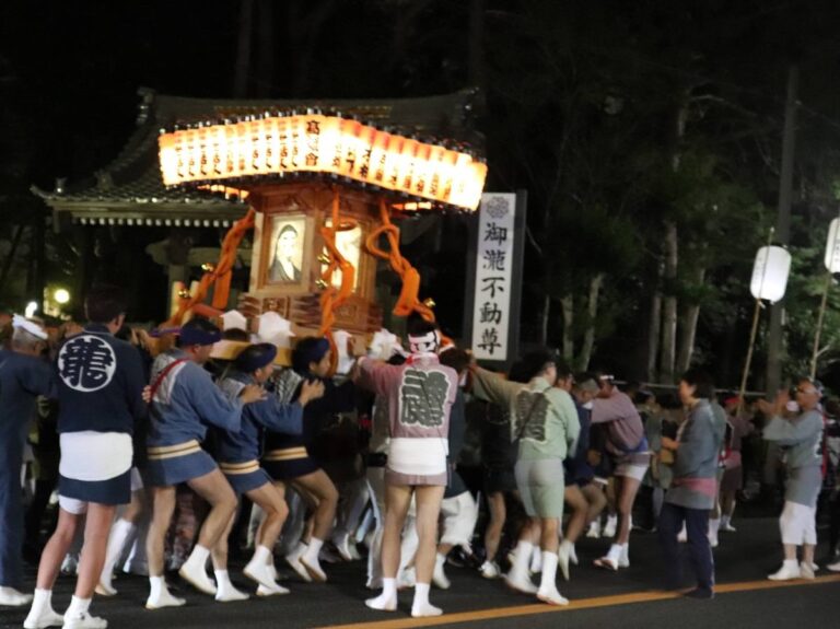 夜、提灯を屋根のように並べた万燈神輿を法被と白足袋姿の担ぎ手が大勢で担いで進む様子。右手に「御瀧不動尊」と書かれた寺号標が立つ