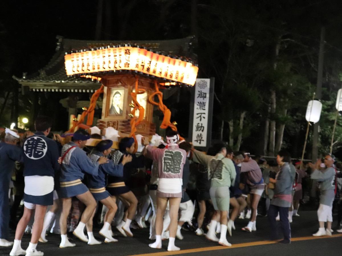 夜、提灯を屋根のように並べた万燈神輿を法被と白足袋姿の担ぎ手が大勢で担いで進む様子。右手に「御瀧不動尊」と書かれた寺号標が立つ