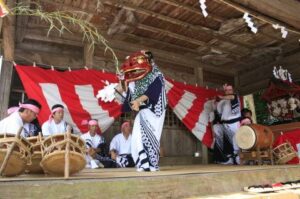 神楽殿の板張りの舞台で、紅白の幕を背に獅子頭をかぶった舞手が舞い、脇に締太鼓と大太鼓の囃子方が並んでいる