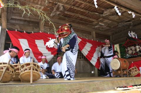 神楽殿の板張りの舞台で、紅白の幕を背に獅子頭をかぶった舞手が舞い、脇に締太鼓と大太鼓の囃子方が並んでいる