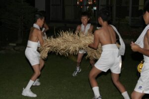 夜、豆絞りの手ぬぐいを肩にかけ白い短パン姿の少年たちが、藁を束ねて編んだ太い綱を数人がかりで抱え、芝生の上を進んでいる。背後は明かりの灯った建物のガラス窓