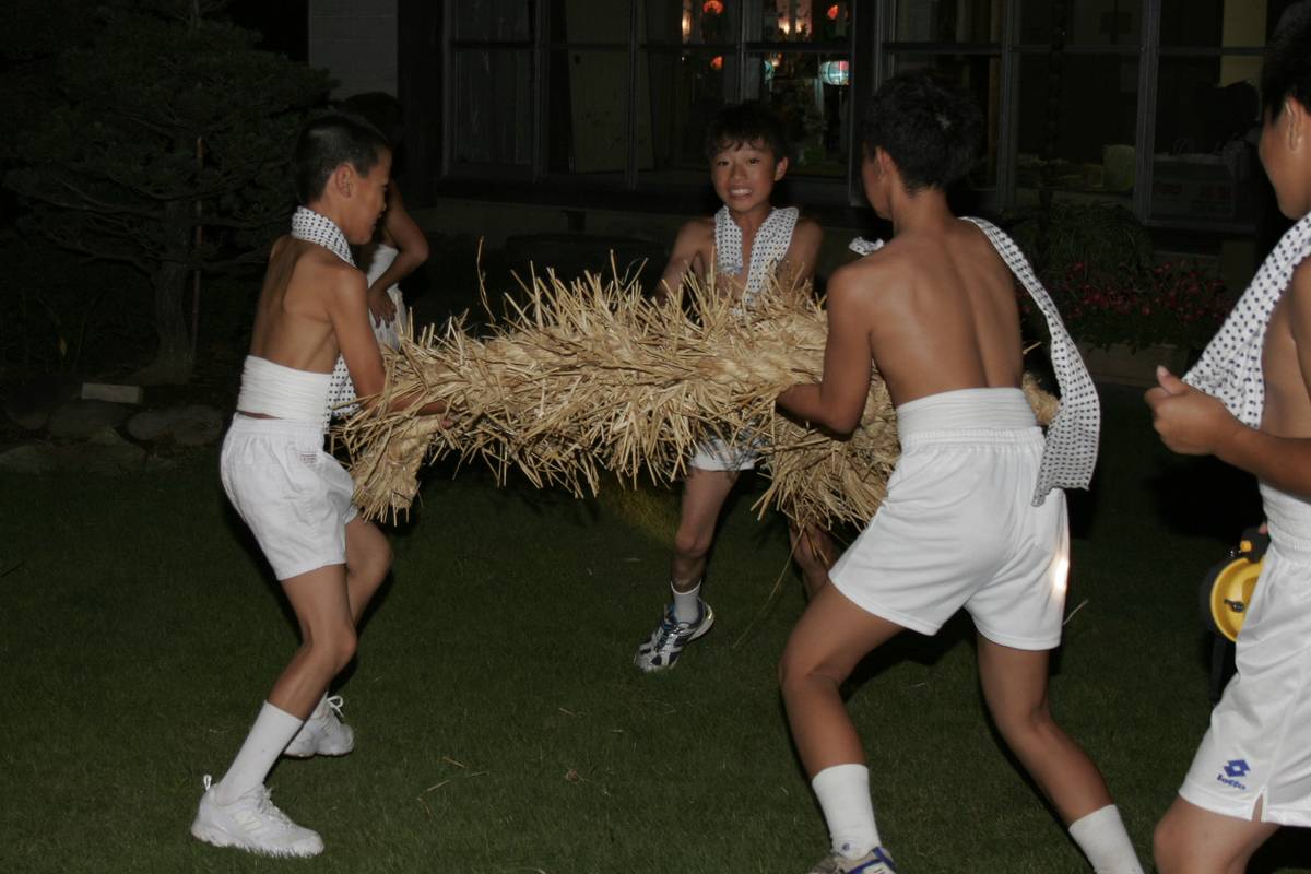 夜、豆絞りの手ぬぐいを肩にかけ白い短パン姿の少年たちが、藁を束ねて編んだ太い綱を数人がかりで抱え、芝生の上を進んでいる。背後は明かりの灯った建物のガラス窓