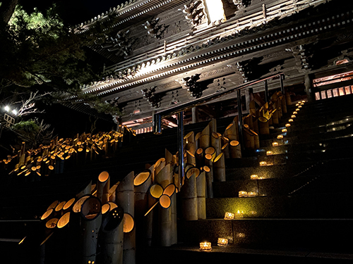 夜の誕生寺で、斜めに切った青竹の灯籠が数十本まとまって内側から橙色に光り、右奥へ伸びる石段の上にも小さな灯りが点々と置かれ、上部にライトアップされた仁王門の軒下の組物がせり出している