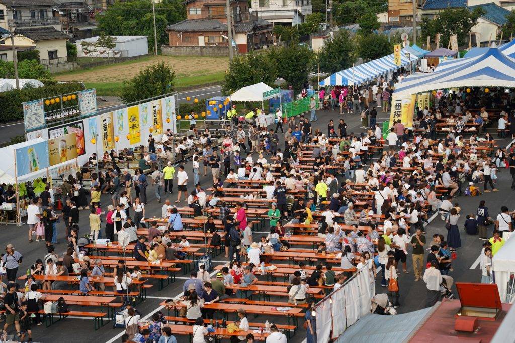町役場の駐車場一面にオレンジ色のビアテーブルとベンチが並び、大勢の来場者が着席している。奥に青と白のストライプの屋台テントとビールブースの列