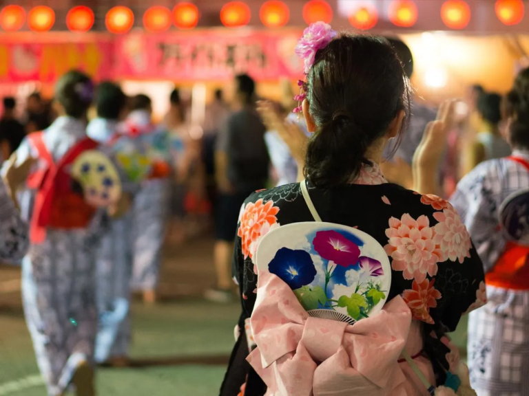 朝顔柄のうちわを帯にさした浴衣姿の女性の後ろ姿と、赤提灯の下で踊る盆踊りの輪