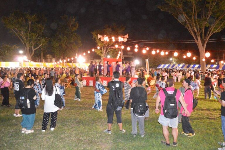 夜のけやきの森公園。中央のやぐらの舞台で浴衣や紫の法被の踊り手が踊り、放射状に張られた提灯の下を大勢の来場者が囲んでいる