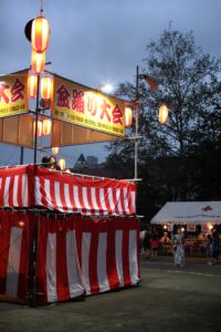 夜の盆踊り大会。提灯を吊るした紅白幕のやぐらと、屋台のテントが並ぶ会場