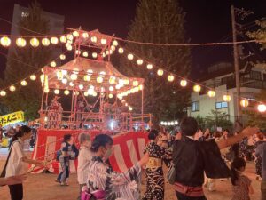 公園の盆踊り。提灯を吊るしたやぐらを囲み、浴衣姿の人たちが輪になって踊っている