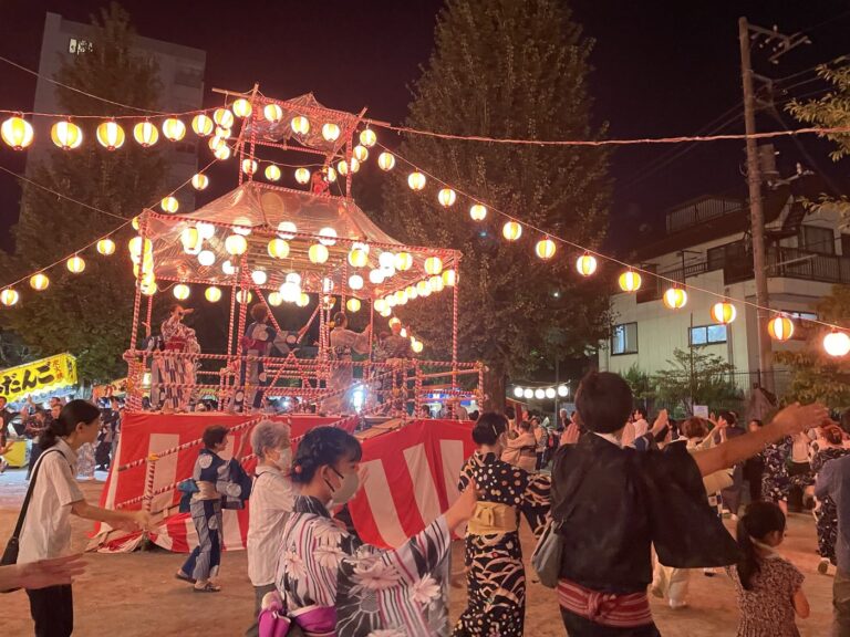公園の盆踊り。提灯を吊るしたやぐらを囲み、浴衣姿の人たちが輪になって踊っている