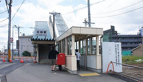 水色の尖った屋根の小さな駅舎に「北柏駅 KITA-KASHIWA STATION」の看板が掲げられ、手前に赤いポストが立つJR常磐線北柏駅の北口