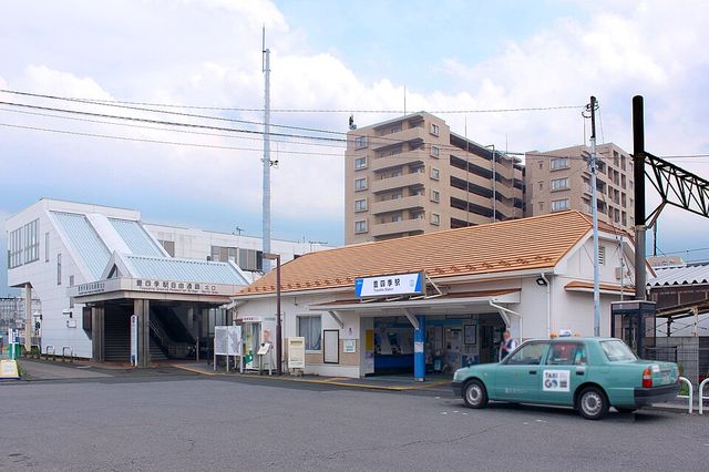 オレンジ色の瓦屋根に「豊四季駅 Toyoshiki Station」の駅名看板を掲げた平屋の駅舎と、白い自由通路の跨線橋