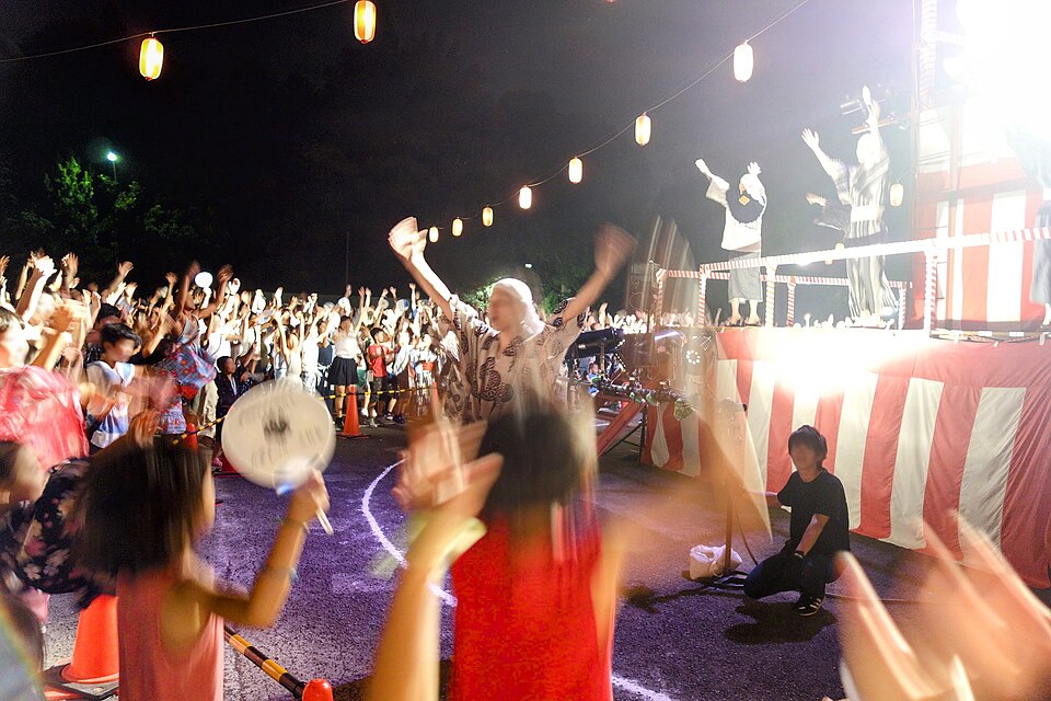 紅白幕を巻いた櫓の前で、提灯の下に大勢の人が集まり手を上げて踊る夜の盆踊り