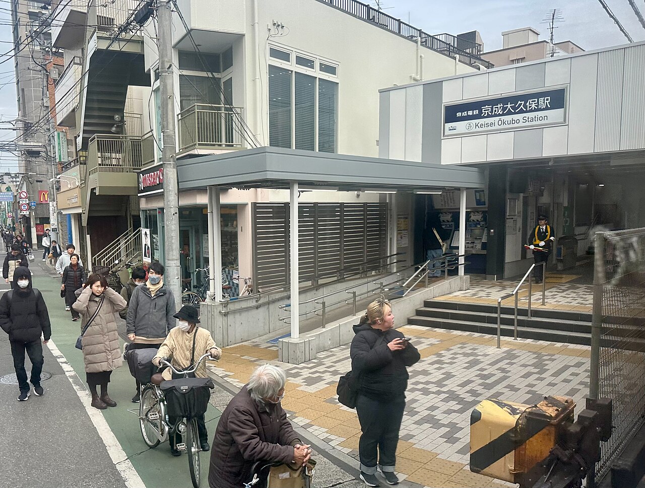 京成大久保駅の駅舎出入口と、その前を歩く人や自転車