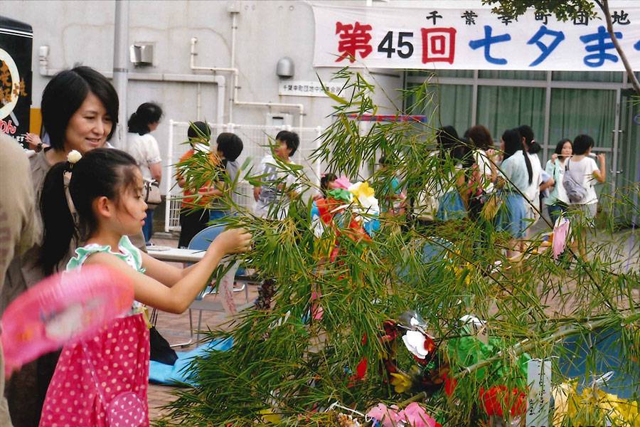 千葉幸町団地の七夕まつり。集会所の壁に「第45回七夕まつり」の横断幕が張られ、笹飾りに短冊を結ぶ親子が写る