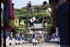 鵜原の大名行列で毛槍や沙熊を掲げて進む青い法被姿の男衆と、後方の神輿・社殿