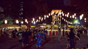夜の会場で紅白幕を巻いたやぐらと提灯の下、浴衣姿の人が輪になって踊る盆踊り